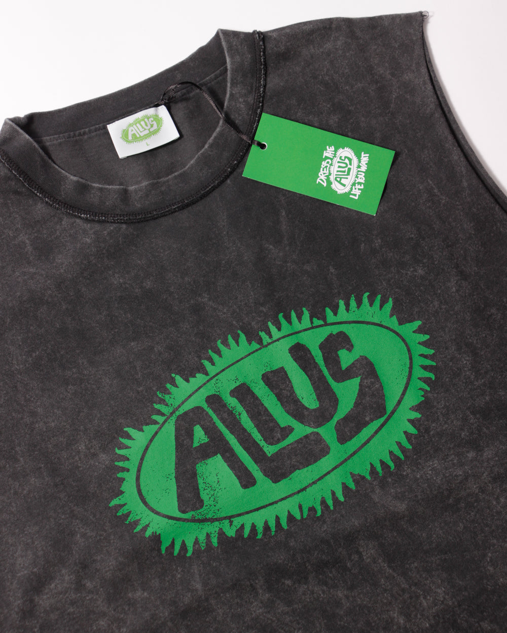 Close-up of the ALIUS sleeveless washed black tank top featuring a bold green logo on the chest and branded green hangtag with "Dress the life you want" slogan.