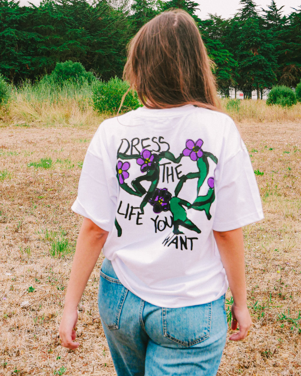 Back view of a woman wearing the white ALIUS "Wild Bloom" t-shirt with purple flowers and the phrase "Dress the life you want", standing in a natural field setting.