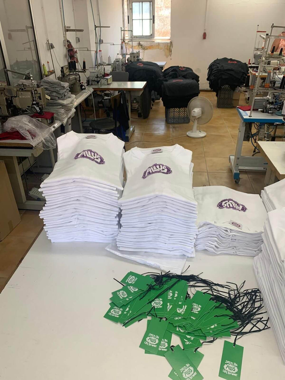 Stacks of white and black ALIUS branded tank tops folded neatly in a local textile workshop, with green hang tags reading "Zero is the ALIUS – Life is What You Make It" displayed in the foreground. Sewing machines and production setup visible in the background.