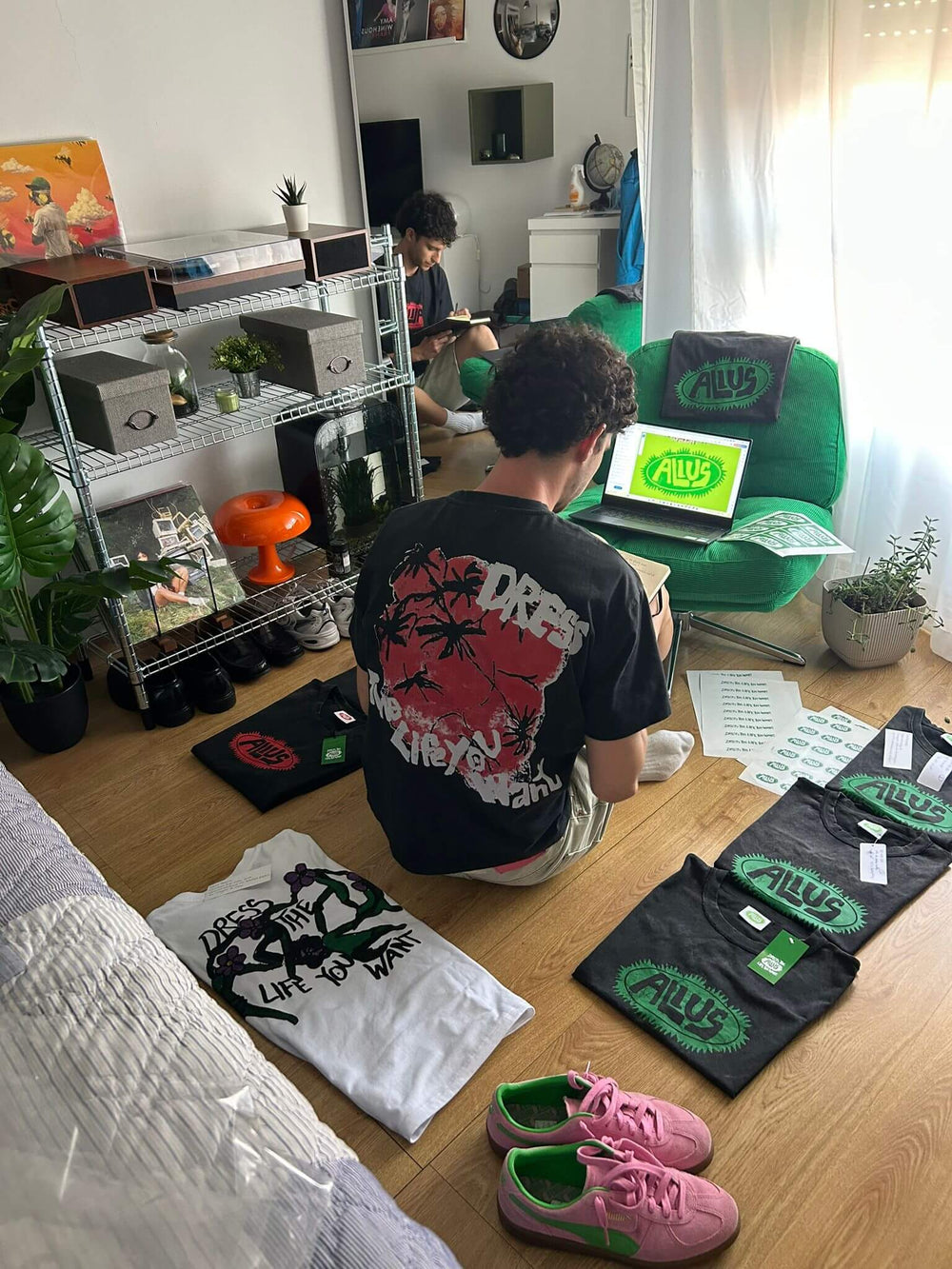 Two young men working on ALIUS clothing in a modern, cozy room. One wears a black t-shirt with the message 'Dress the Life You Want', surrounded by branded ALIUS shirts, a laptop showing the logo, and pink and green Puma sneakers. Urban streetwear and creative studio atmosphere.