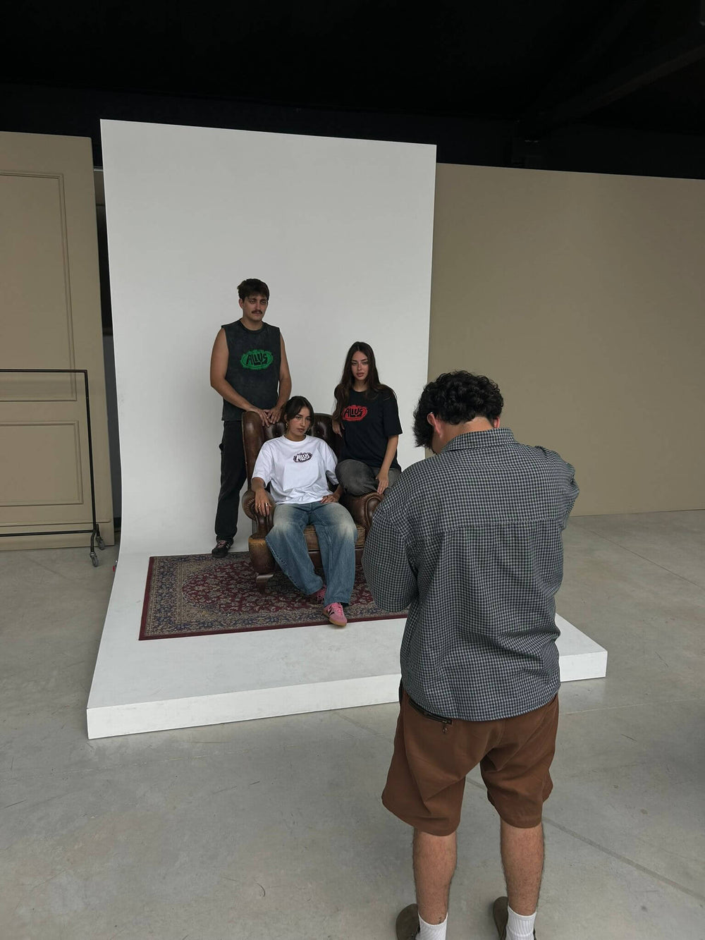 Behind-the-scenes of an ALIUS photoshoot featuring three models wearing branded t-shirts in black, green, and white with the ALIUS logo. The models pose in front of a white backdrop and vintage rug, while a photographer captures the scene in a minimalist studio setting.