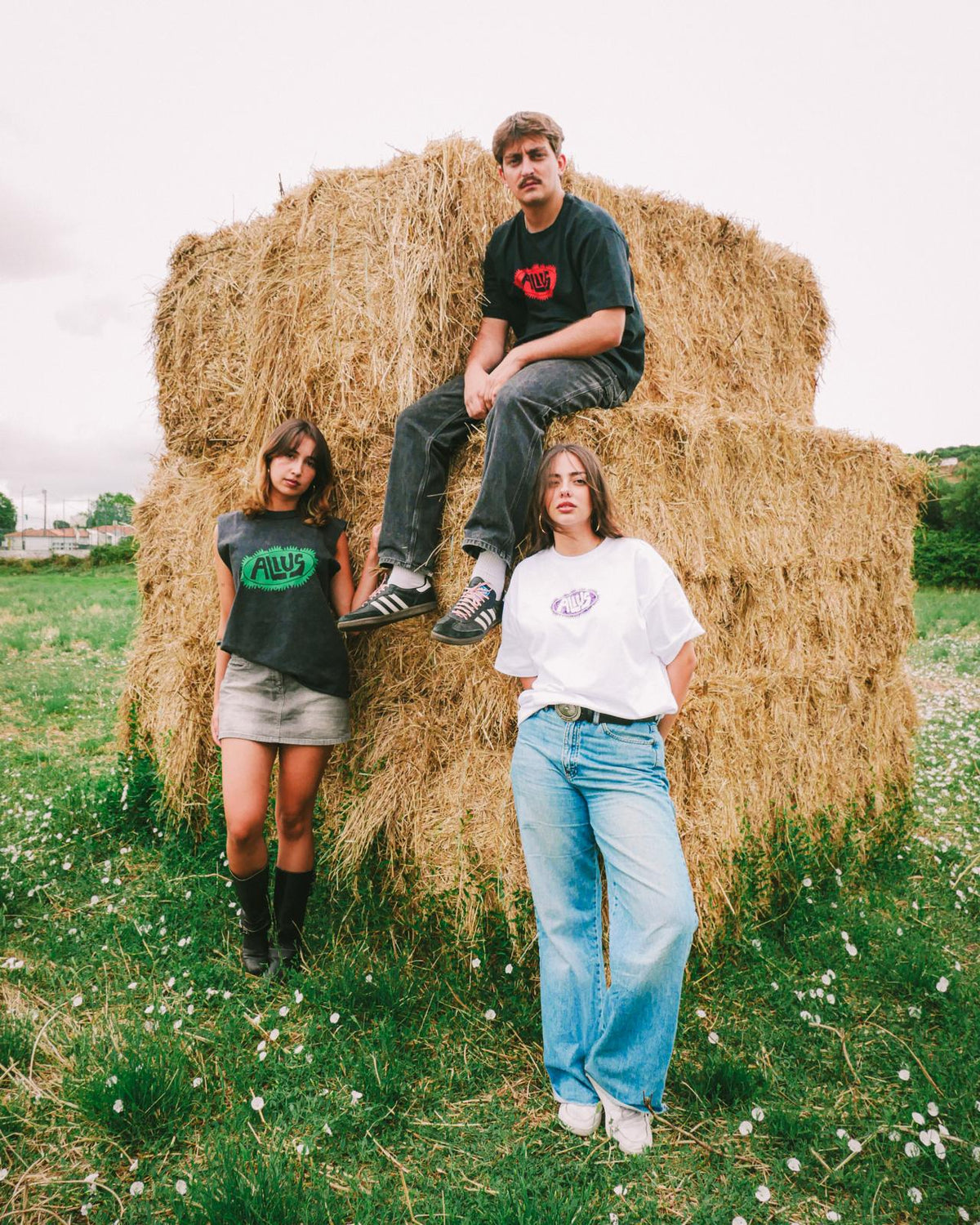 Three models wearing ALIUS streetwear t-shirts posing in a countryside setting with hay bales, showcasing unisex minimalist fashion with bold graphic prints.