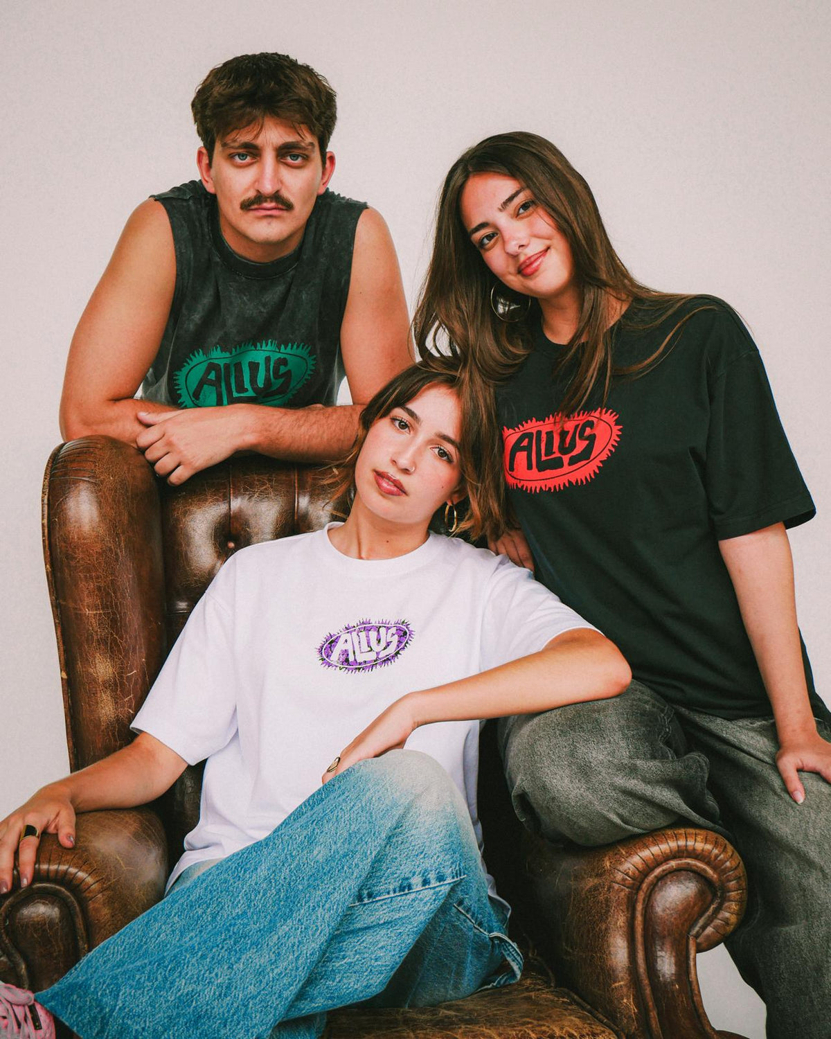 Three young models wearing ALIUS streetwear graphic tees in a studio setting, sitting on a vintage leather chair. Unisex fashion with bold colors and minimalist logo designs.